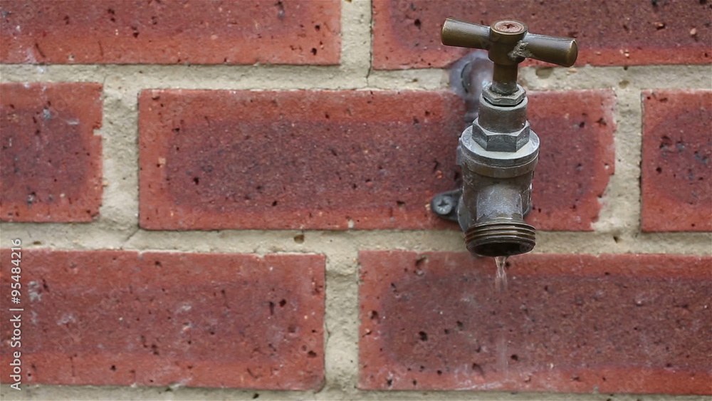 Outside tap leaking water drips against a bright red brick wall Stock
