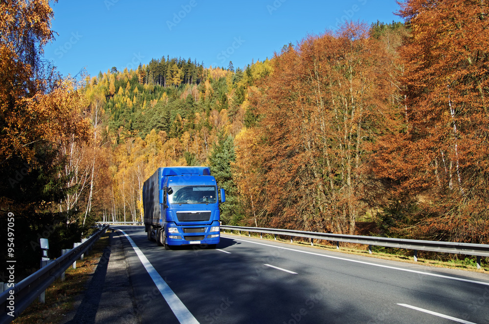 Blue truck on asphalt road in a wooded valley below the mountain, blazing with autumn colors. Sunny autumn day with blue sky.