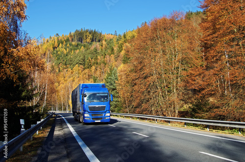 Obraz na płótnie Blue truck on asphalt road in a wooded valley below the mountain, blazing with autumn colors. Sunny autumn day with blue sky. w salonie