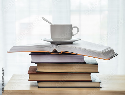 Stack of books and a cup with a spoon on a wooden table against the window.