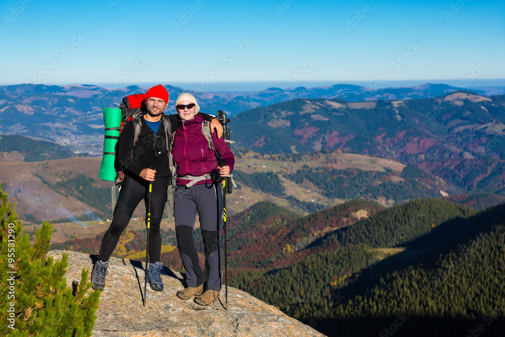 Male and Female Hikers with Backpacks Sporty Clothing and Trekking Poles Staying on High Rock with Mountain View with Autumnal Colors Forest and Blue Sky on Background