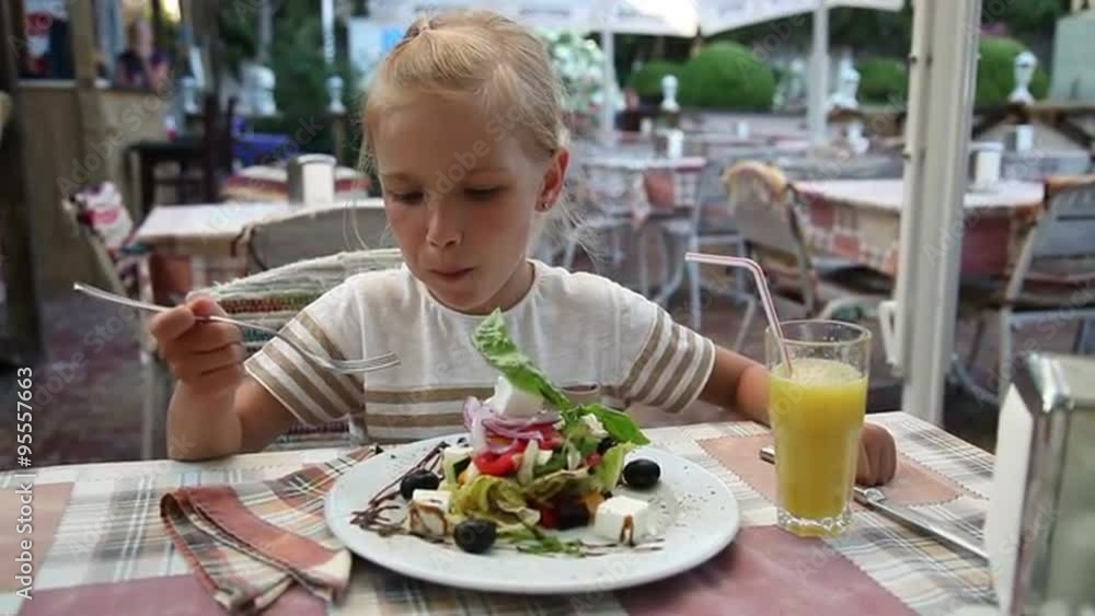 Little girl eating a Greek salad in the cafe and looking at the camera Little girl eating a Greek salad in the cafe and looking at the camera
