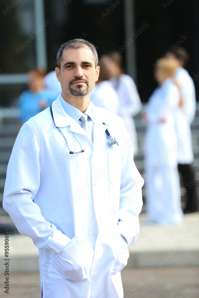 Fototapeta premium Happy doctor with medical stuff behind standing against clinic entrance