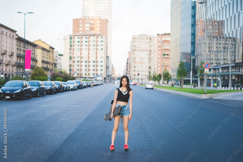 Young handsome asiatic long brown straight hair woman skater posing in the middle of the street in the city, looking in camera, pensive - thinking future, serious, thoughtful concept