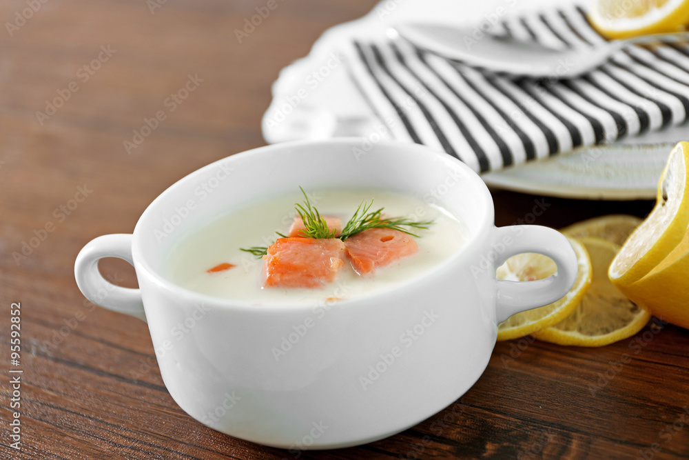 Delicious salmon cream soup in white bowl with lemon and striped cotton serviette on wooden table, close up Delicious salmon cream soup in white bowl with lemon and striped cotton serviette on wooden table, close up