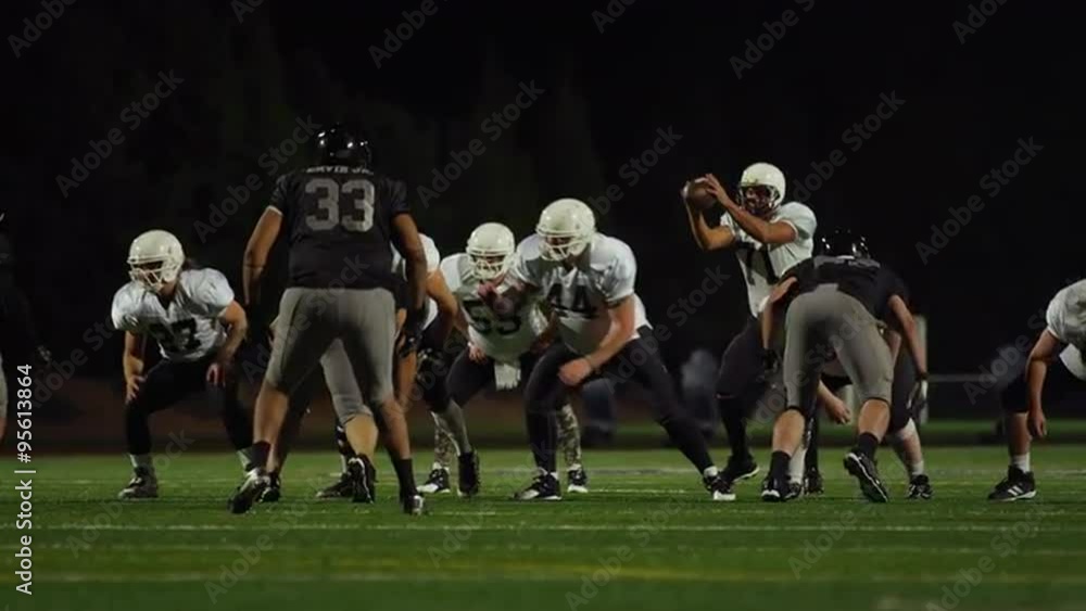 A football is snapped during a game and then thrown toward the camera to a teammate at night