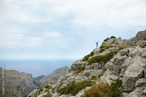 Young man standing on cliff with hands raised on top of mountain at Sa Colobra, Majorca, Spain. Happy man winner on top of mountain.
