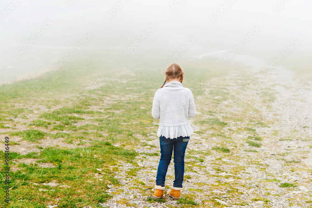 Cute little girl of 8 years old playing outdoors on a very foggy day, wearing grey warm pullover, back view