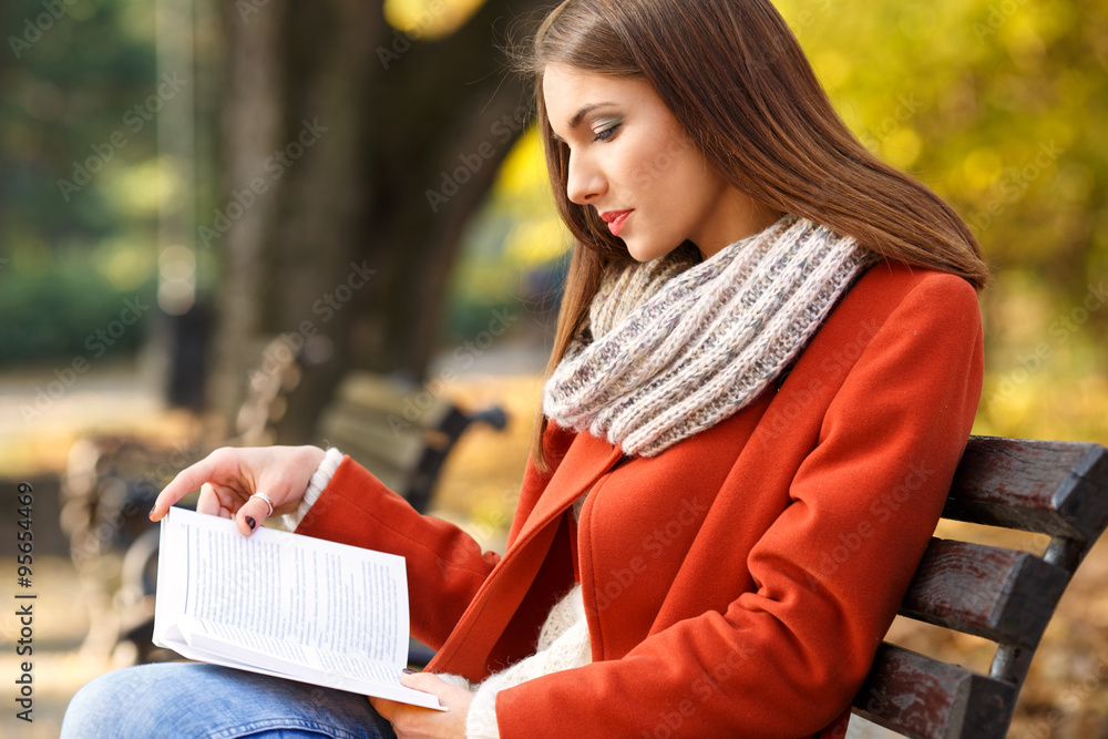 Fototapeta premium Young girl sitting on a park bench and reading a book, on a beautiful autumn day