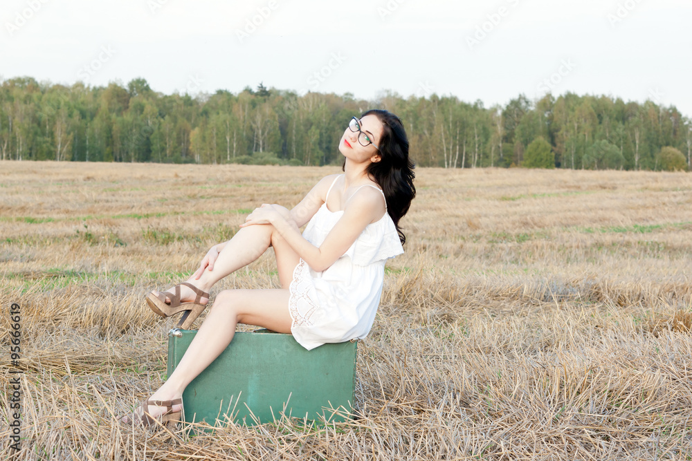 girl in a short dress sitting on an old suitcase on a background of beveled fields