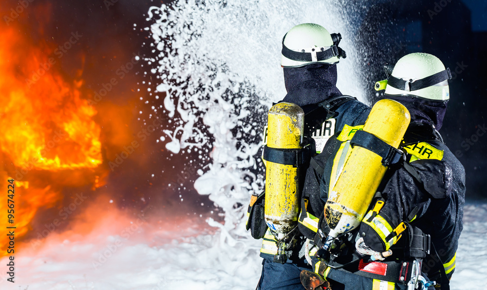 Obraz premium Feuerwehr - Feuerwehrmänner löschen ein großes Feuer, sie stehen vor einer Feuerwand und tragen Schutzkleidung
