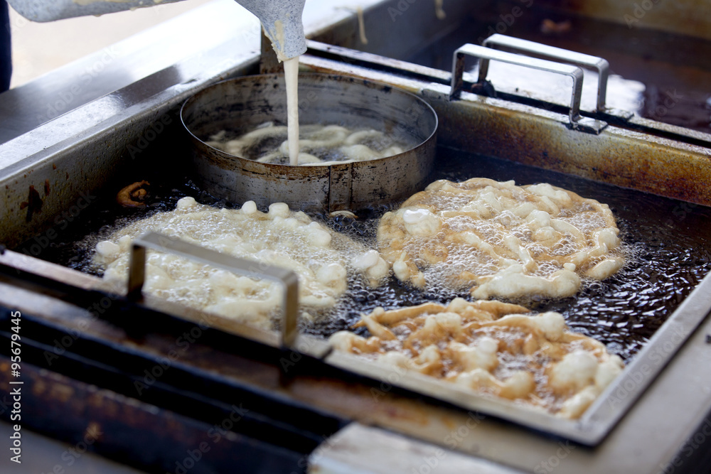 Inside the funnel cake booth, vendor is pouring funnelcake batter though a funnel into boiling hot oil to make a popular fair/festival dessert.
