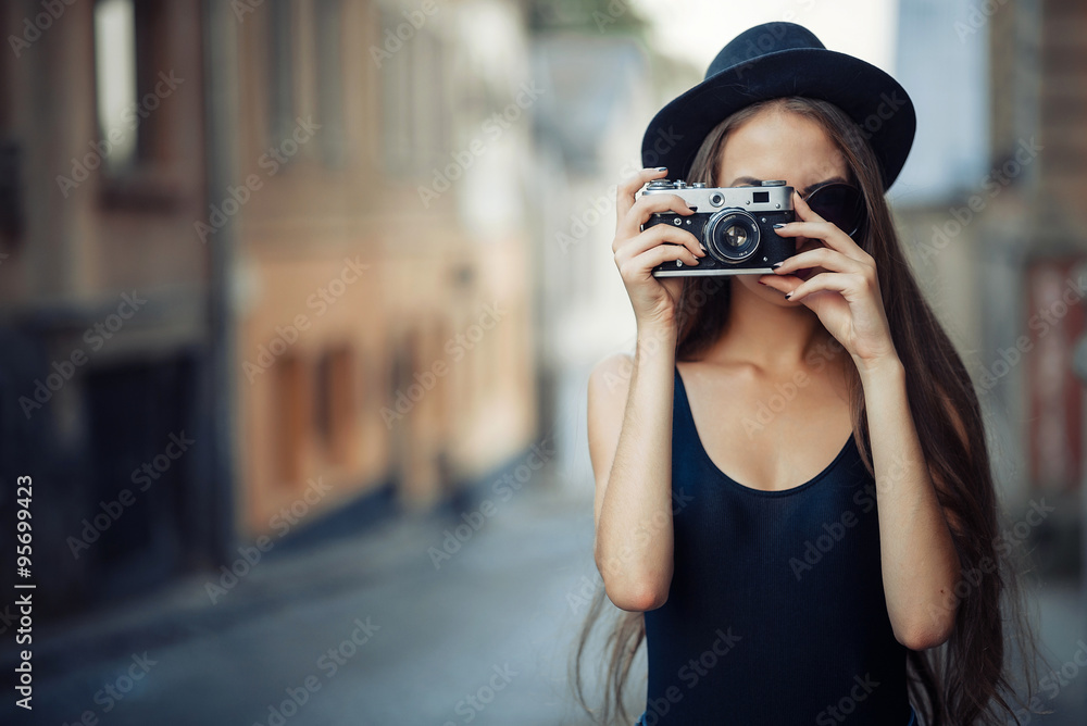 young beautiful brunette girl in a hat, sunglasses, t-shirt and jeans with a film camera in hand walks along the ancient city