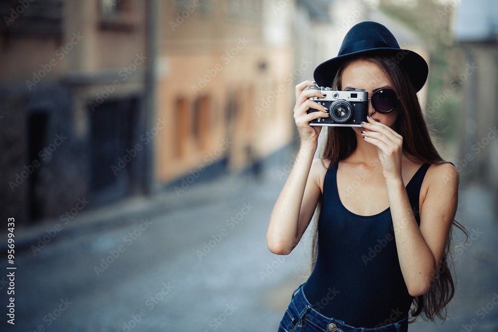 young beautiful brunette girl in a hat, sunglasses, t-shirt and jeans with a film camera in hand walks along the ancient city