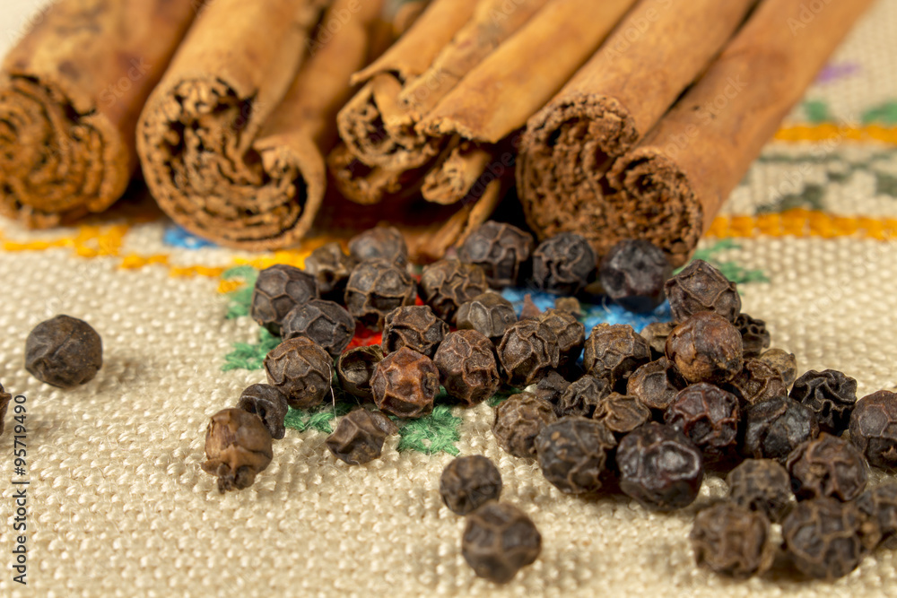 Obraz premium Grains of black pepper and several cinnamon sticks over a embroidered cloth