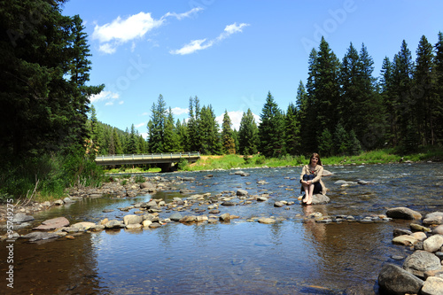 Alone with her thoughts, young woman relaxes and puts her barefeet in the Gallatin River in the Gallatin River Valley in Montana.  Bridge sits in background. photo