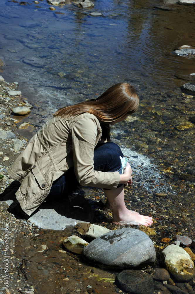 Naklejka premium Young woman sits barefoot besides the Gallatin River in the Gallatin Valley of Montana. She is picking up rocks from the riverbed.