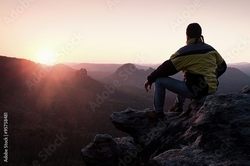 Rear view of male hiker in yellow black jacket sitting on rocky cliff while enjoying daybreak above valley