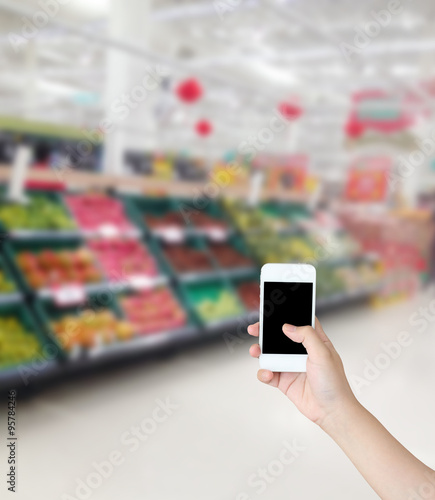 Hand holding mobile phone with Vegetables and fruit on shelf in