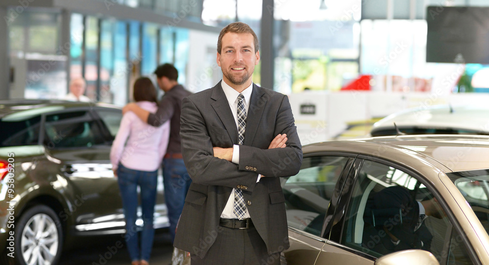 erfolgreicher Geschäftsmann im Autohandel - im Hintergrund Kunden & Interieur des Autohauses // 
successful businessman in the car trade - in the background customer & interior of the car dealership