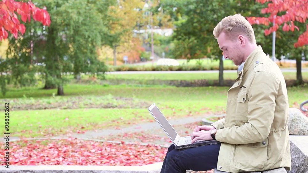 A handsome man in his 20s sitting down on a bench outside with the great colors of autumn behind, holding a laptop computer. A handsome man in his 20s sitting down on a bench outside with the great colors of autumn behind, holding a laptop computer.