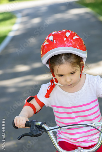 Beautiful little girl with colourful red safety helmet, riding a bicycle in summer park outdoors