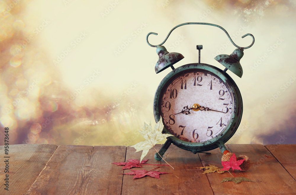 image of vintage alarm clock next to autumn leaves on wooden table in front  of abstract blurred background. retro filtered Photos | Adobe Stock