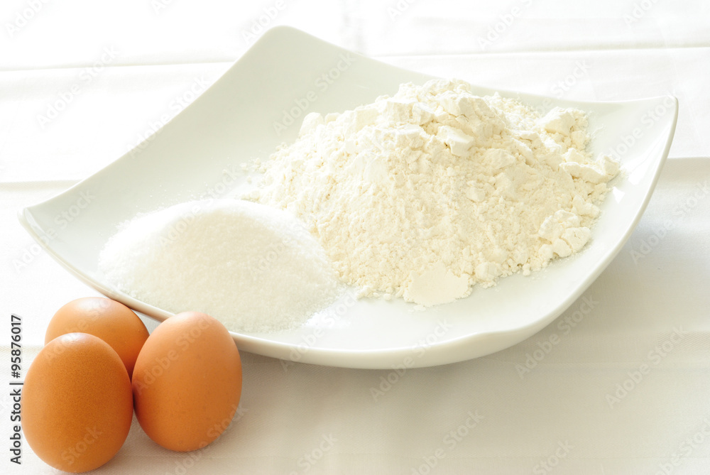 farina, zucchero, uova, ingredienti per deliziosi dolci, torte e cupcakes, sistemati in un piatto quadrato sopra una tovaglia bianca farina, zucchero, uova, ingredienti per deliziosi dolci, torte e cupcakes, sistemati in un piatto quadrato sopra una tovaglia bianca