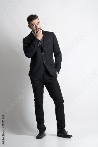 Skeptical man in suit with hand on his chin looking at camera. Full body length portrait over gray studio background. 