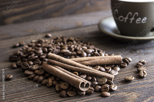 Coffee beans and cinnamon sticks bark lie on a wooden table in front of blurred cup of coffee. Blur, focus on near-stick #95887479