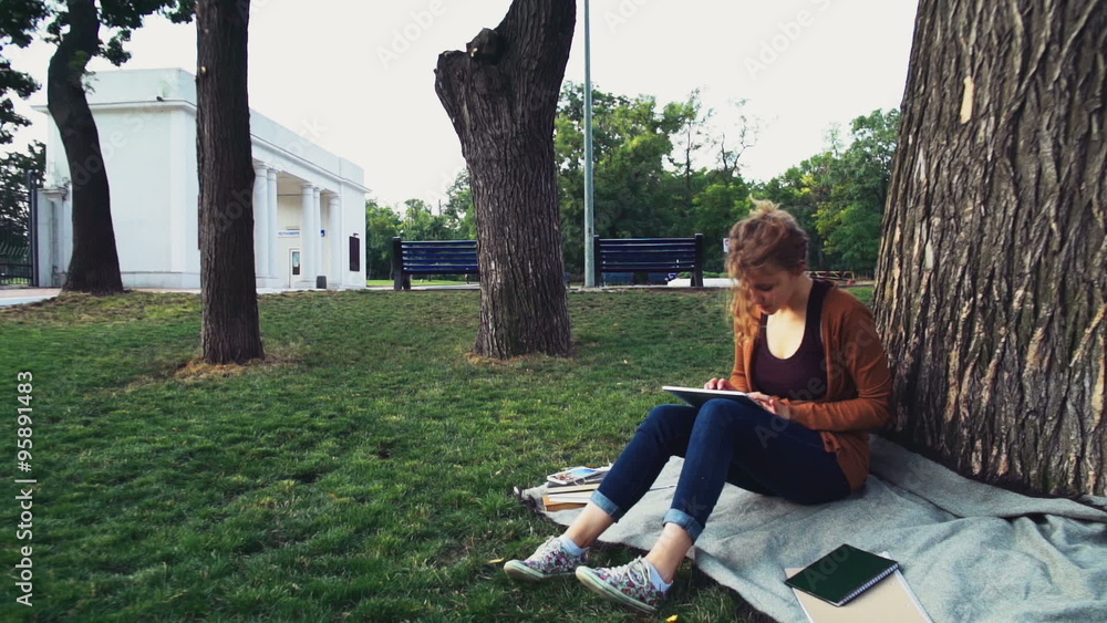 female student sitting under a tree in park and using tablet female student sitting under a tree in park and using tablet
