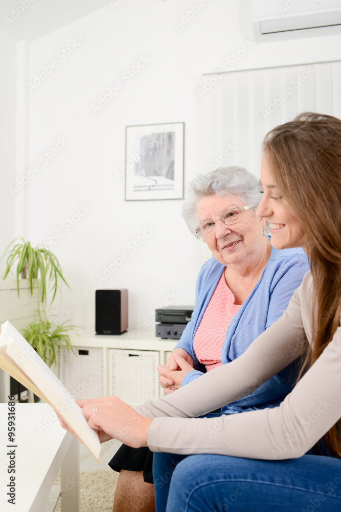 cheerful young woman reading book for old senior woman at home cheerful young woman reading book for old senior woman at home