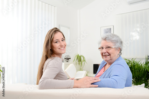 cheerful young woman reading book for old senior woman at home