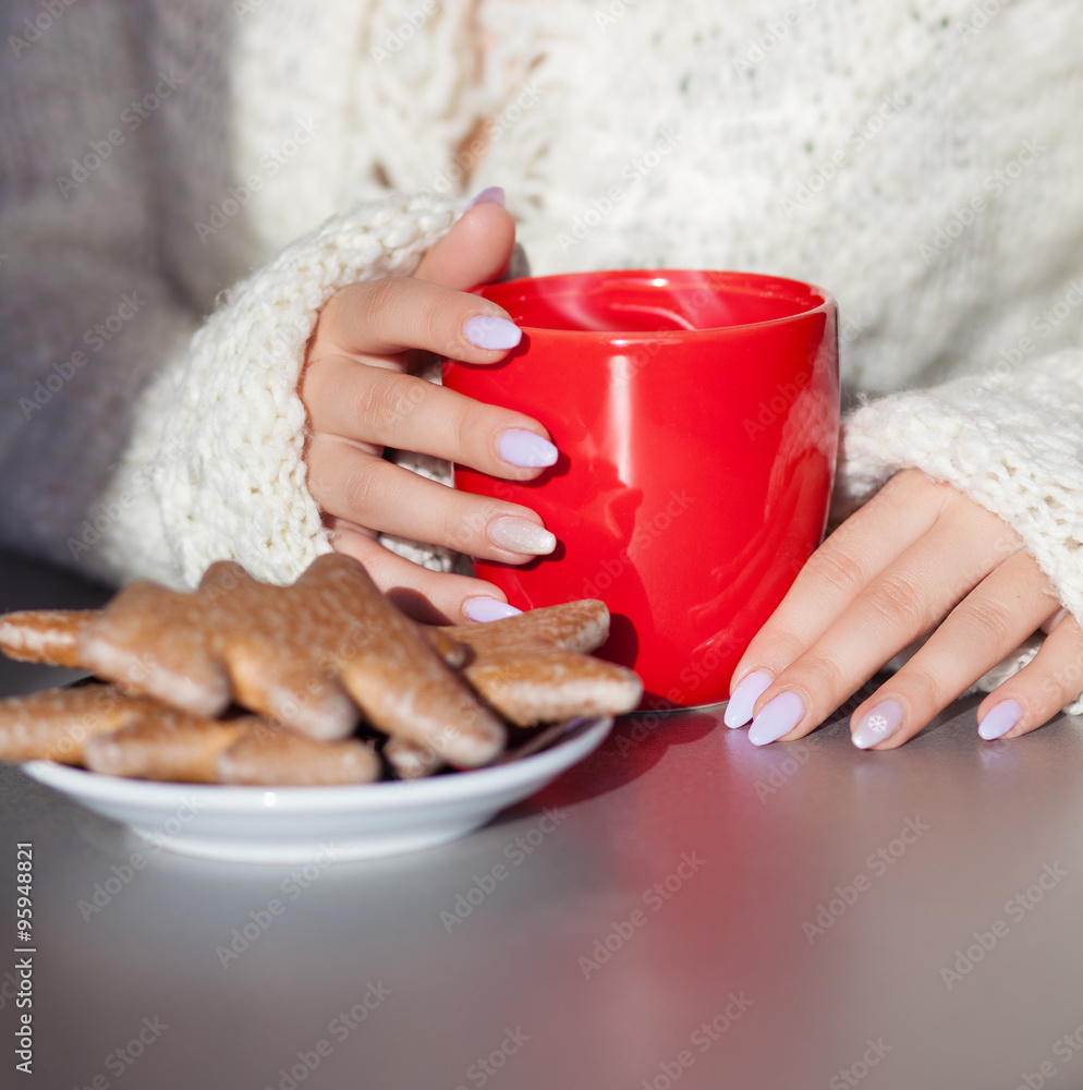 Close up of woman's hands holding cup of hot coffee drink. She is wearing warm cardigan. Christmas gingerbread on the table. Winter chill out and lifestyle concept. Close up of woman's hands holding cup of hot coffee drink. She is wearing warm cardigan. Christmas gingerbread on the table. Winter chill out and lifestyle concept.
