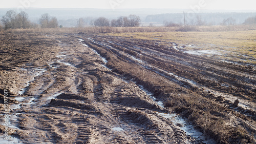 Vanishing dirty road through fallow field with mud and puddles at early spring
