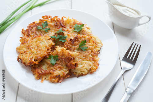 Fried potato pancakes or latke traditional homemade Hanukkah celebration food with greens and sour cream sauce recipe in plate on rustic white wooden table background. Natural organic meal