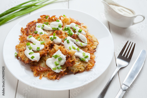 Homemade fried potato pancakes or latke traditiona Hanukkah celebration food with greens and sour cream sauce served in dish on white rustic wooden table background