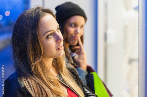 Two women friends in the evening just after sunset are doing shopping in around the city. They stopped in front of a shop window that illuminates their faces. One of them looks aghast article © loreanto