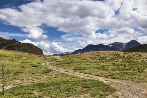 Winding dirt road among green meadows going to snow peaks