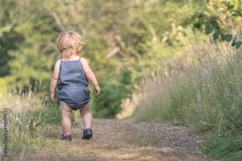 Blond baby in blue jeans costume walking away down on green path, baby's first steps