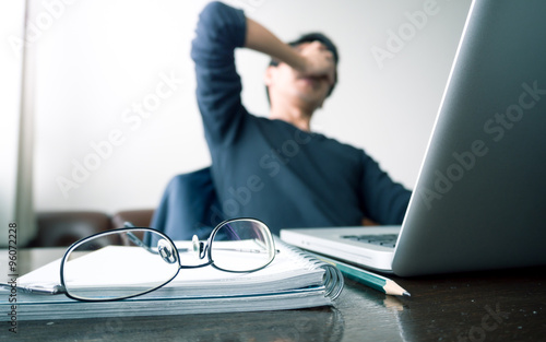 man with his hand holding his face taking a brake from working with laptop computer and notebook with eye glasses on wooden desk. concept of stress/rest/tension/failed/discourage. green filtered.