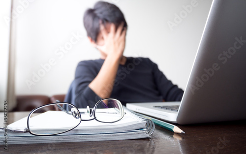 man with his hand holding his face taking a brake from working with laptop computer and notebook with eye glasses on wooden desk. concept of stress/rest/tension/failed/discourage