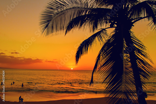 Tropical island sunset with silhouette of palm trees, hot summer day vacation background, golden sky with sun setting over horizon, people swimming in the ocean at sunset