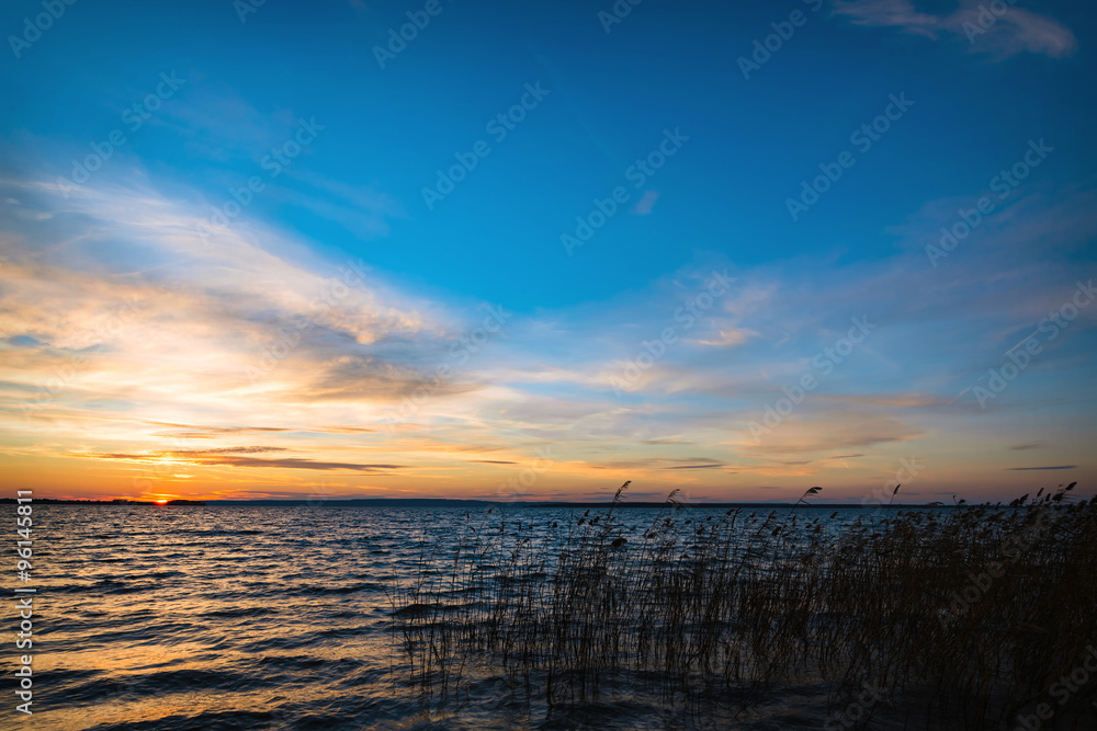 Obraz premium Lake sunset over calm water with reeds, featuring vibrant blue sky, orange horizon light, and gentle ripples, ideal for nature travel background, eco banners, and serene landscape design