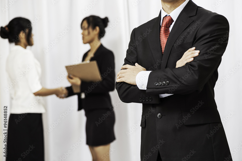 businessman stand folding his arms while two women shake hands in the background
businessman stand folding his arms while two women shake hands in the background