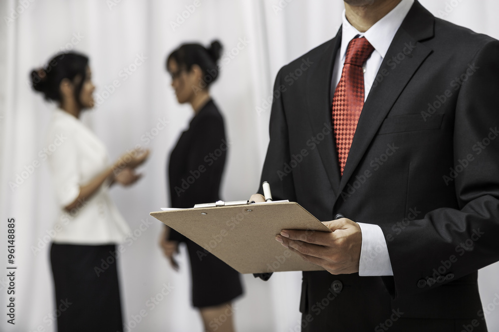 businessman holding his notepad while two women are talking in the background
businessman holding his notepad while two women are talking in the background