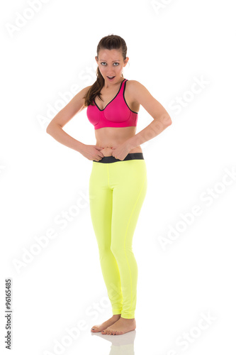 Brunette woman in sports neon yellow leggings and pink bra with shocked and surprised expression shows excess of fat on belly on white background. © anatmari