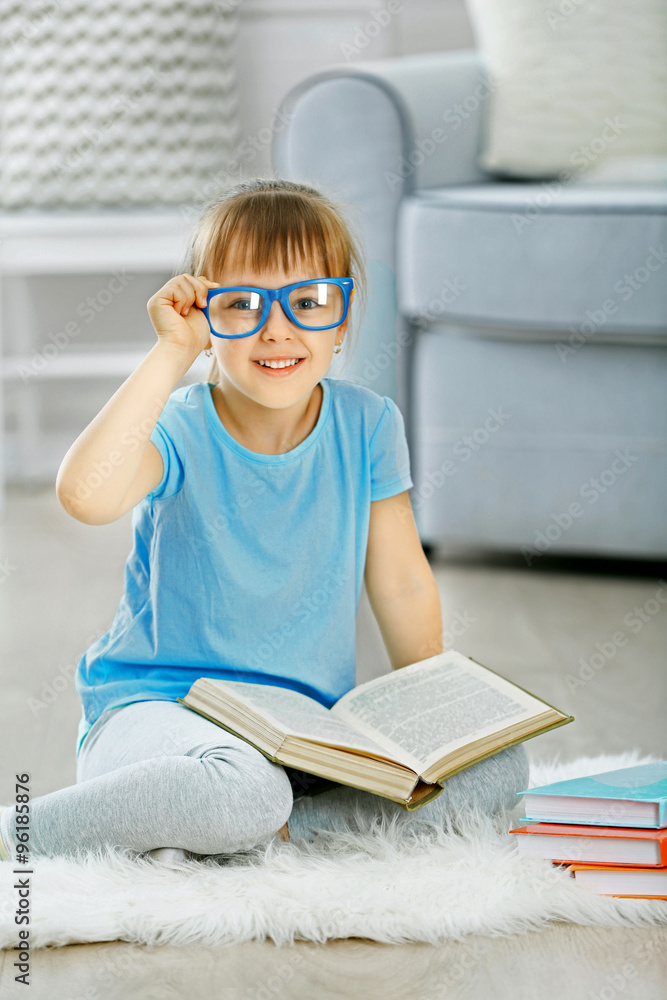 Beautiful little girl sitting on carpet near sofa with book, on home interior background Beautiful little girl sitting on carpet near sofa with book, on home interior background