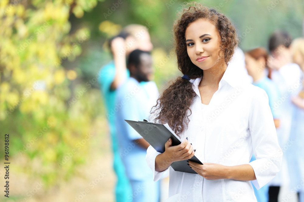 Attractive young woman doctor with clipboard in hands against group of medics, outdoors Attractive young woman doctor with clipboard in hands against group of medics, outdoors