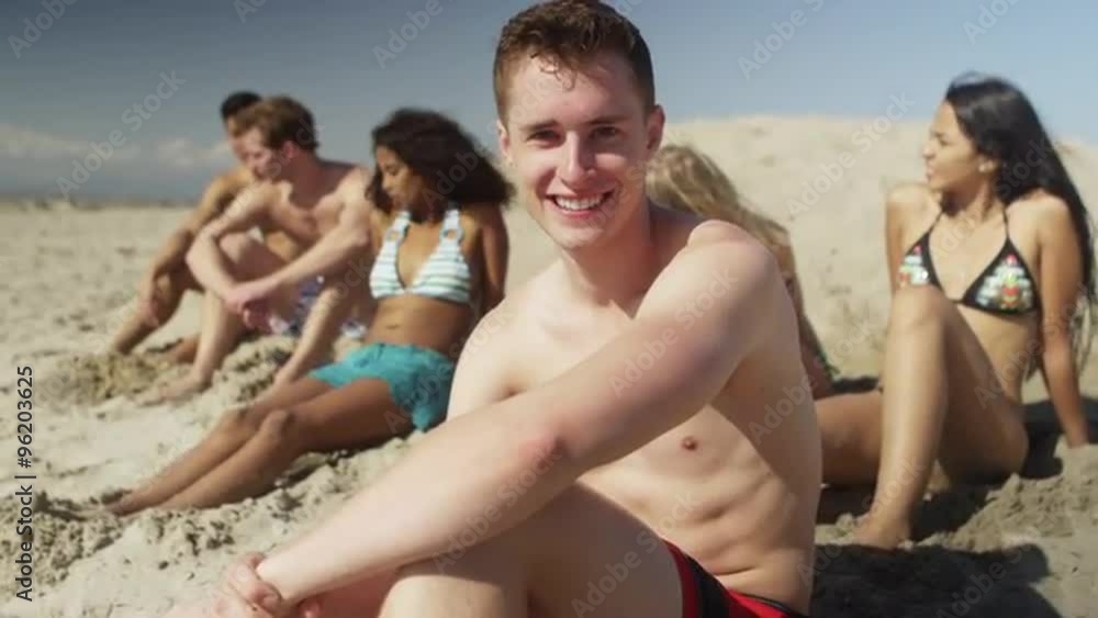 Portrait of young caucasian man sitting in sand with friends on the beach smiling Portrait of young caucasian man sitting in sand with friends on the beach smiling